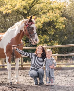 A brown and white paint horse stands with people in a fenced paddock on an autumn day.