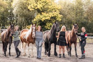 A group of people stand with their horses in a fall setting with golden leaves and trees in the background.