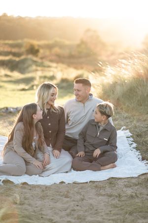 A family sits together on a white blanket at sunset in the dunes, sharing a warm moment together.