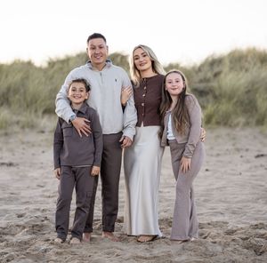 Family wearing coordinated neutral outfits poses together on sandy beach dunes at sunset.