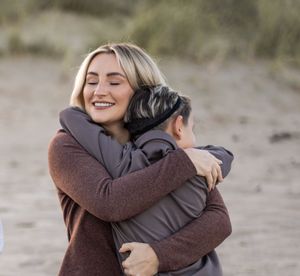 Two people share a warm embrace outdoors on a cloudy day, one wearing a brown sweater and the other in gray.