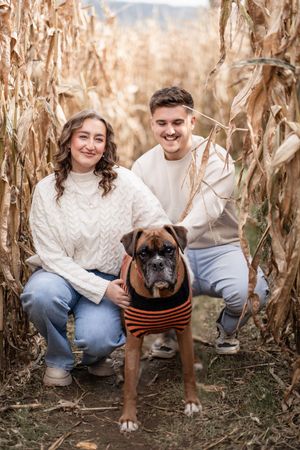 A couple in cozy sweaters poses with their boxer dog wearing a striped shirt in a corn maze.