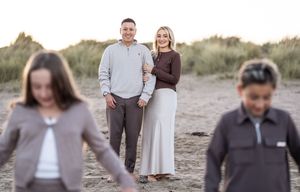 Family members embrace and laugh together while standing on coastal sand dunes in muted tones.
