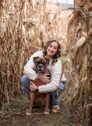 A boxer dog and companion pose together in a cornfield during autumn with dried corn stalks in the background.