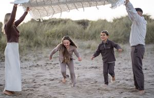 A family plays an energetic game under a blanket being held up like a canopy on the beach.