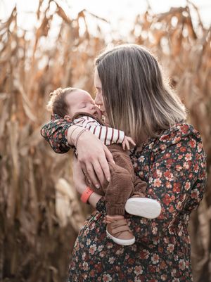 A person in a floral dress holds and cuddles a small brown cat against a backdrop of dried corn stalks.