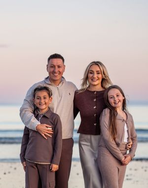 A family of four poses together on the beach at sunset wearing coordinated brown and neutral colored outfits.