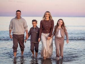 Family wearing coordinated brown and beige outfits walking together in ocean waves at sunset.