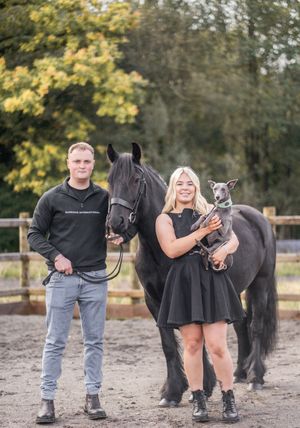 A couple stands with their black horse and small dog in a fenced paddock with autumn trees in the background.