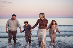 A family holds hands while splashing together in the ocean at sunset with pink skies behind them.