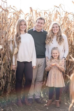 Family poses together in casual attire against a backdrop of dried corn stalks during an autumn photo session.