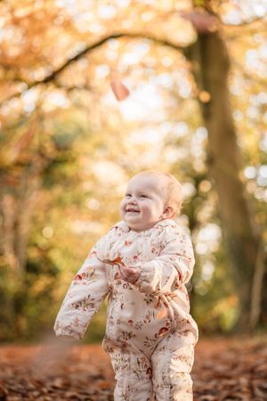 A baby wearing a floral onesie smiles while playing in autumn leaves with golden fall foliage in the background.
