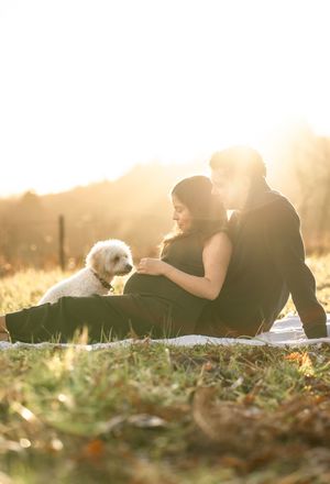 A couple sits with their white fluffy dog on a blanket in a sunlit field during golden hour.