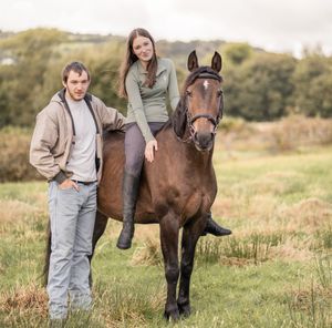 A young child in overalls stands next to a brown horse in a grassy field during autumn.