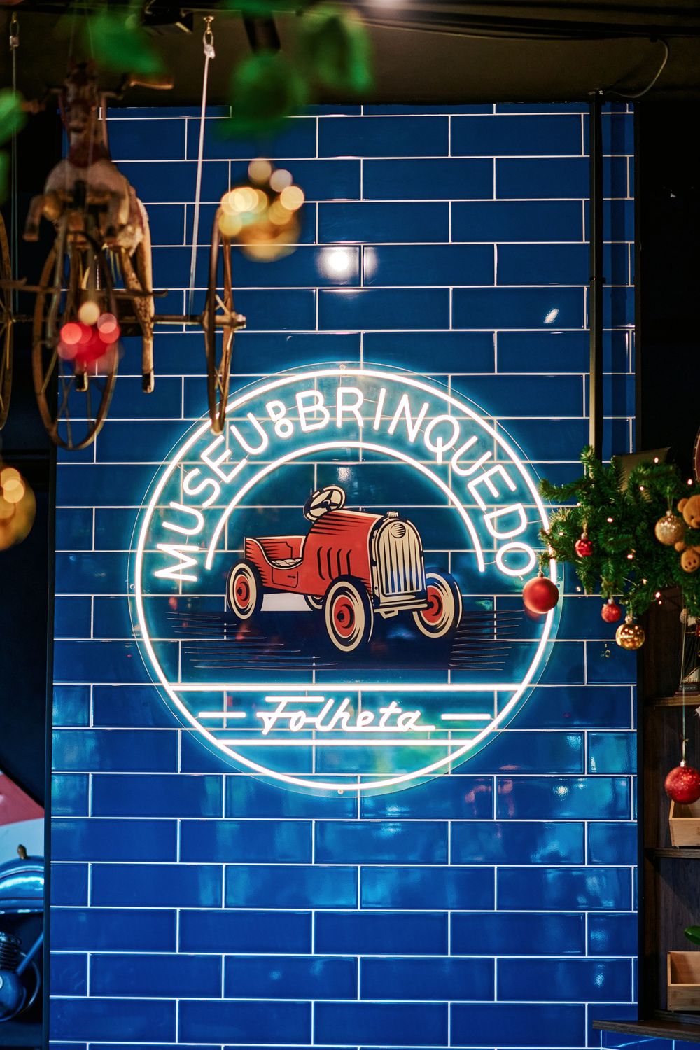 Neon sign for Museu do Brinquedo Folheta on a blue tile wall with Christmas decorations.