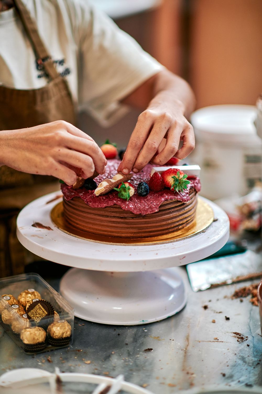 Pastry chef decorating a layered chocolate cake with fresh strawberries and berries on a cake stand.