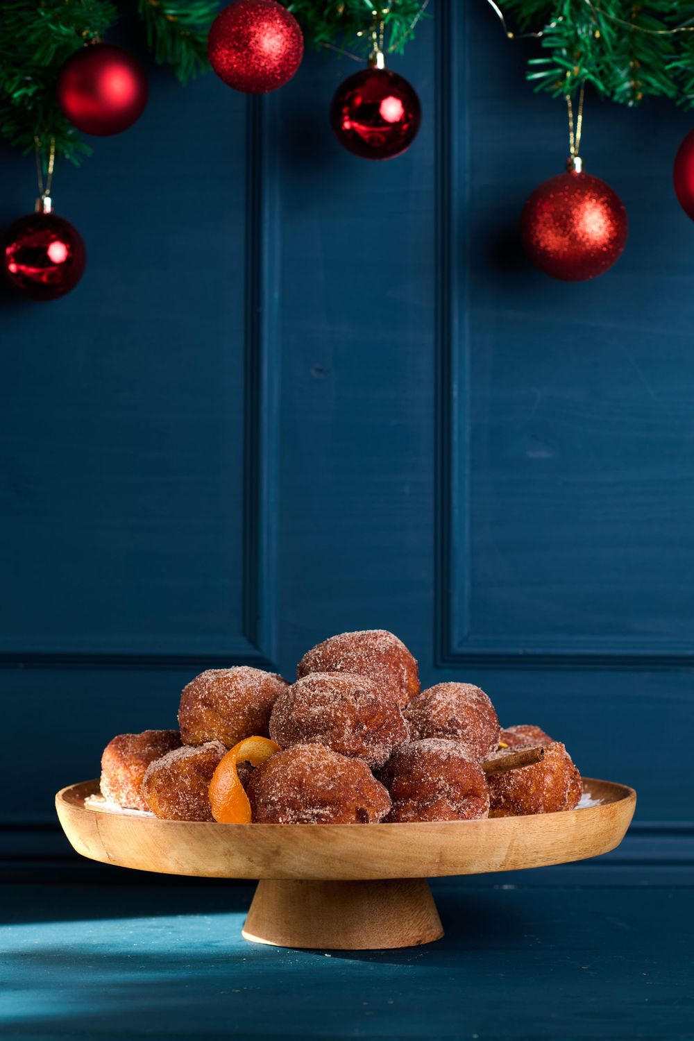 Sugar-dusted donuts piled on a wooden cake stand with Christmas ornaments and greenery in background.