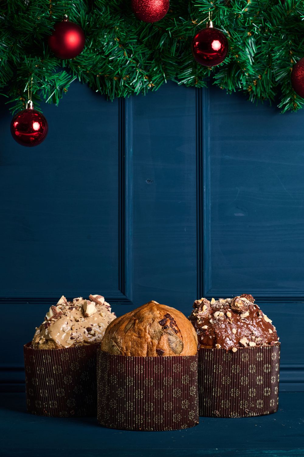 Three artisan panettone breads in decorative packaging displayed under Christmas garland with red baubles.