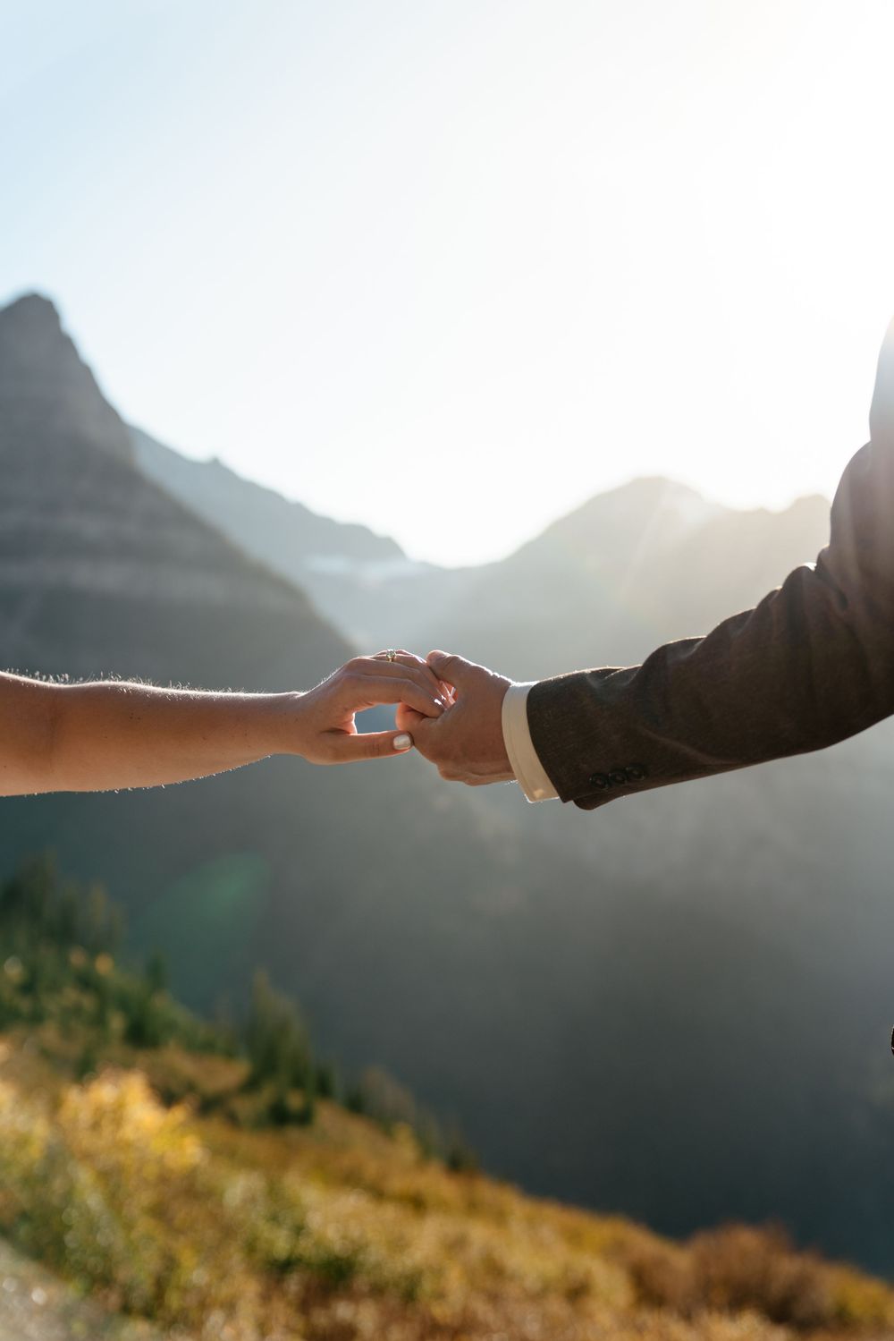 Two hands reach out to touch fingertips against a backdrop of sun-drenched mountains and golden sky at sunset.