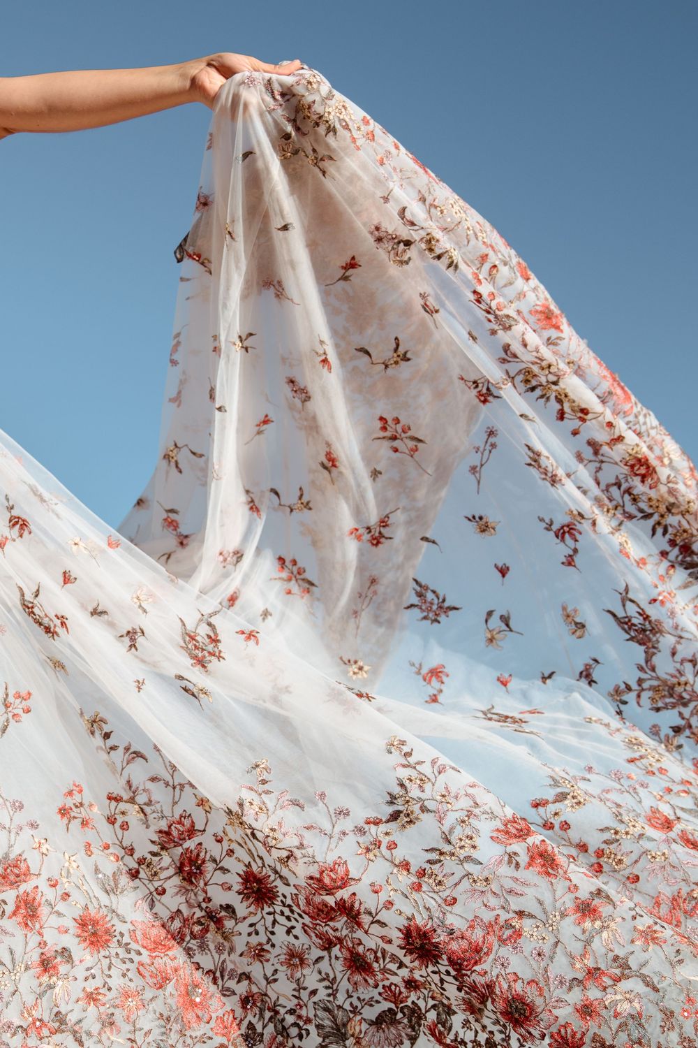 A hand holds up flowing white fabric with delicate red floral patterns against a bright blue sky.