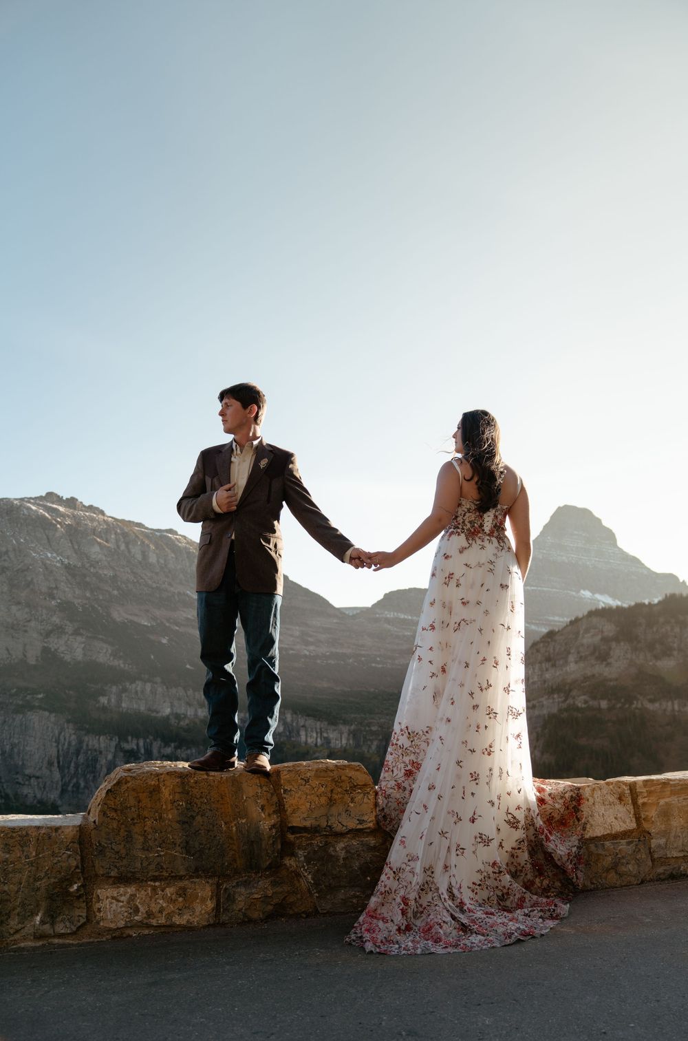 A couple in wedding attire holds hands while standing on a stone wall with mountain peaks silhouetted in the background.