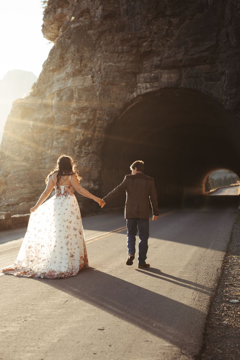 A bride and groom in wedding attire holds hands while walking along a sunlit pathway near stone tunnel at sunset.