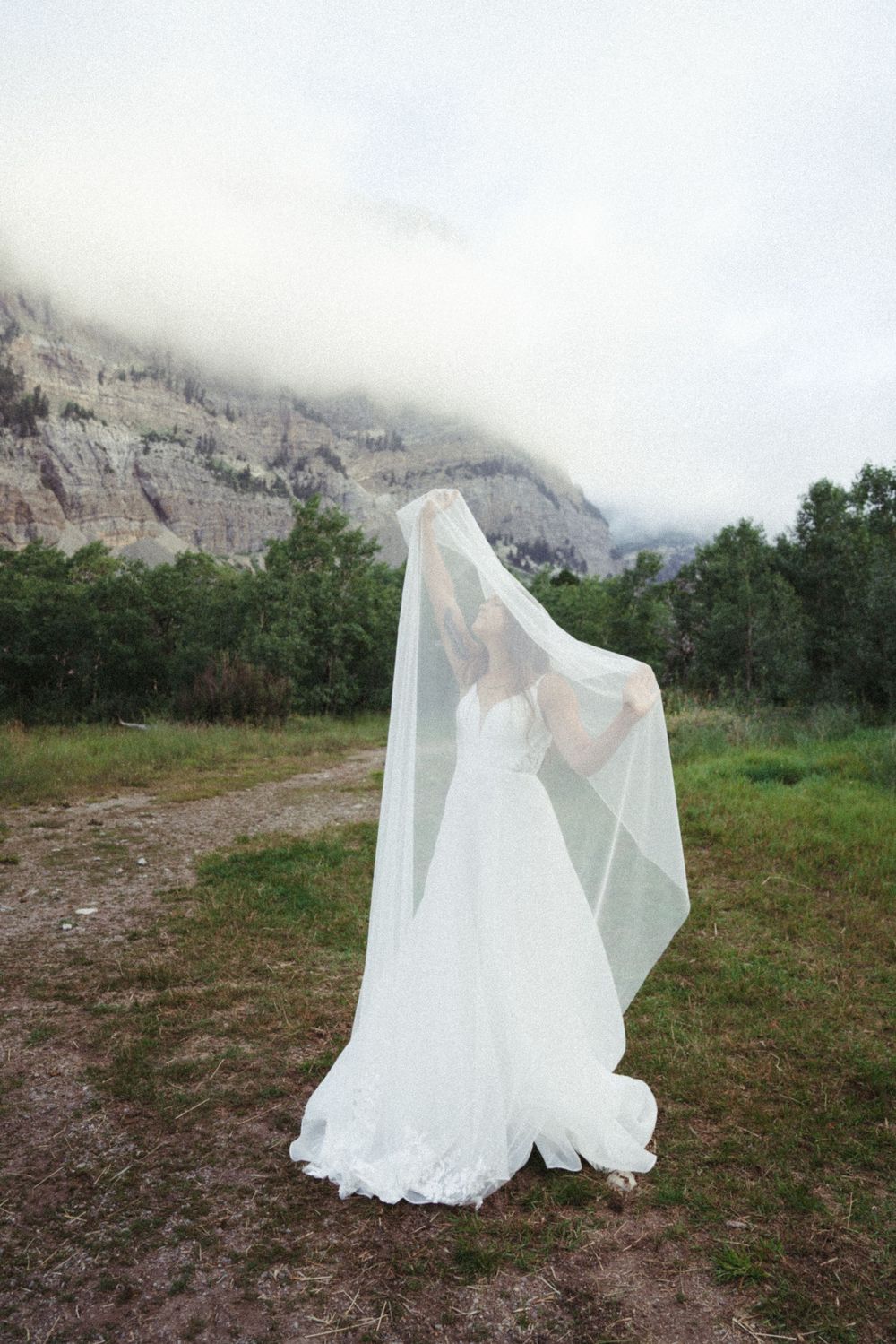 A flowing white wedding dress billows in the wind against a backdrop of misty mountains and green fields.