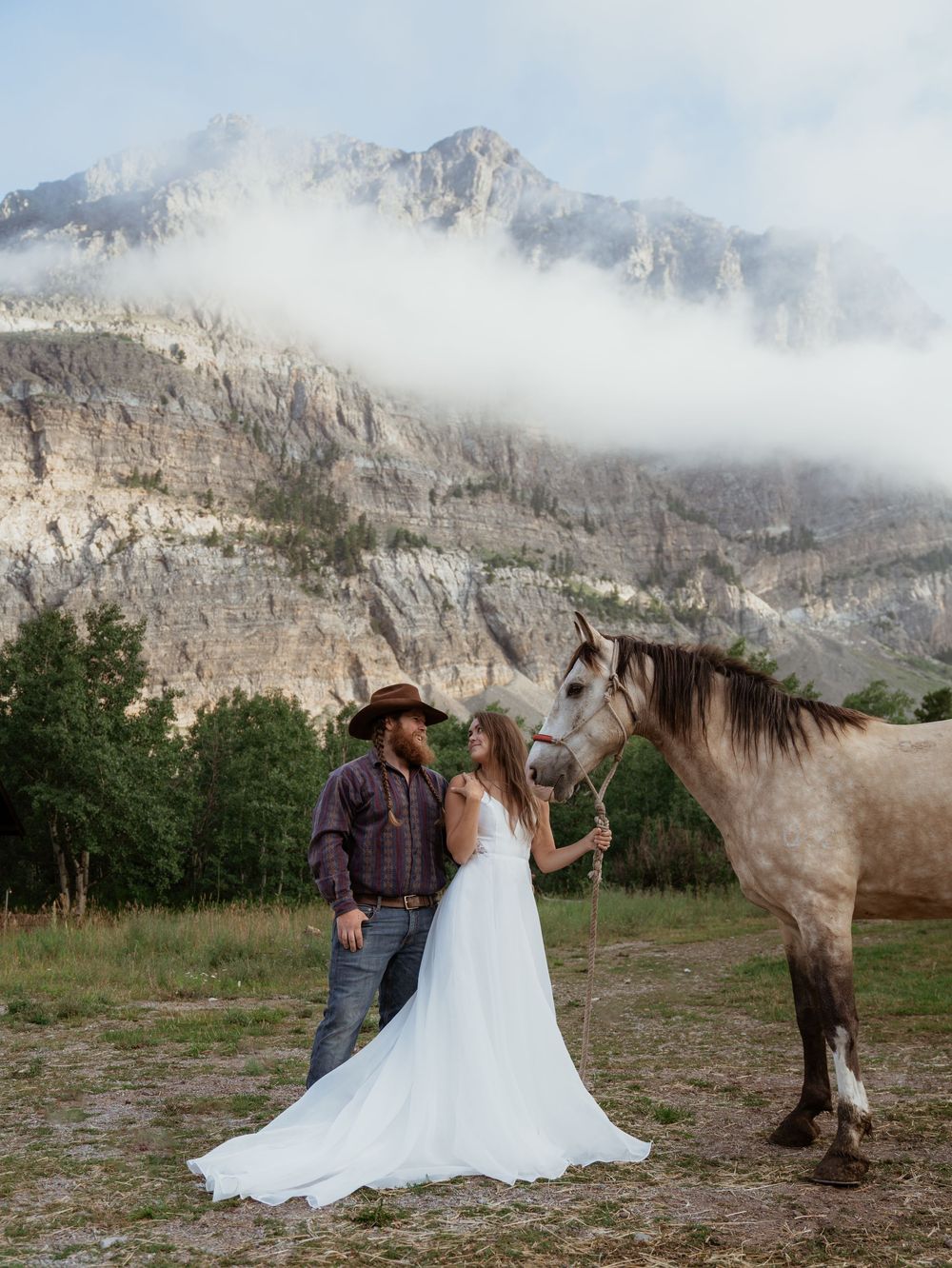 Couple poses with a horse against a backdrop of misty mountains and dramatic cliffs during their wedding photoshoot.