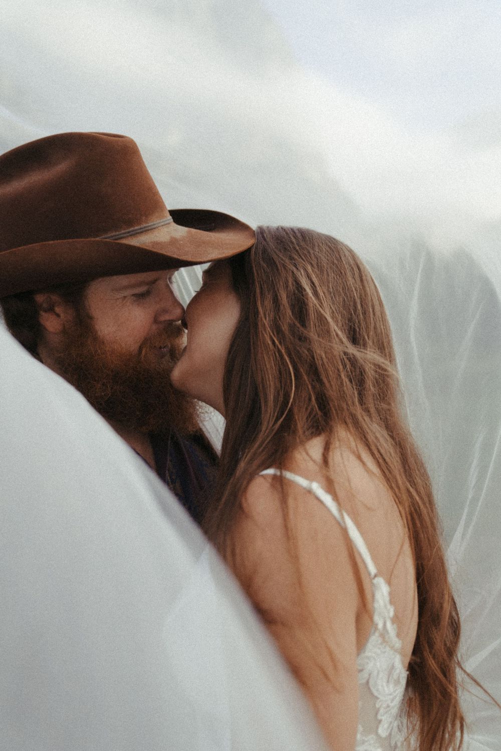A romantic couple kissing under a veil in Glacier National Park, with one wearing a brown cowboy hat and the other in wedding dress.