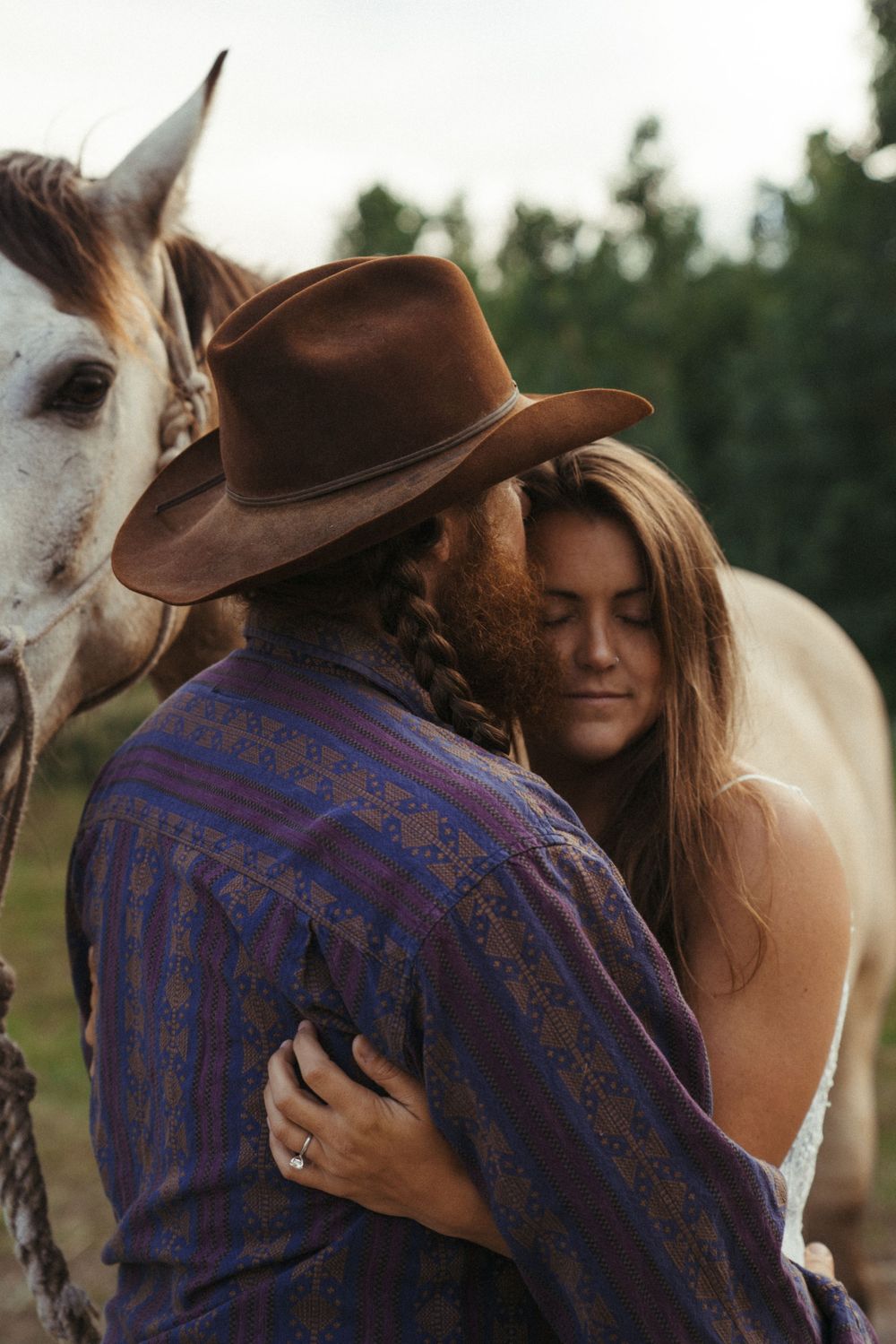 Vintage edit of a groom in a brown cowboy hat and blue patterned shirt shares a tender moment with his bride while horse stands behind them.