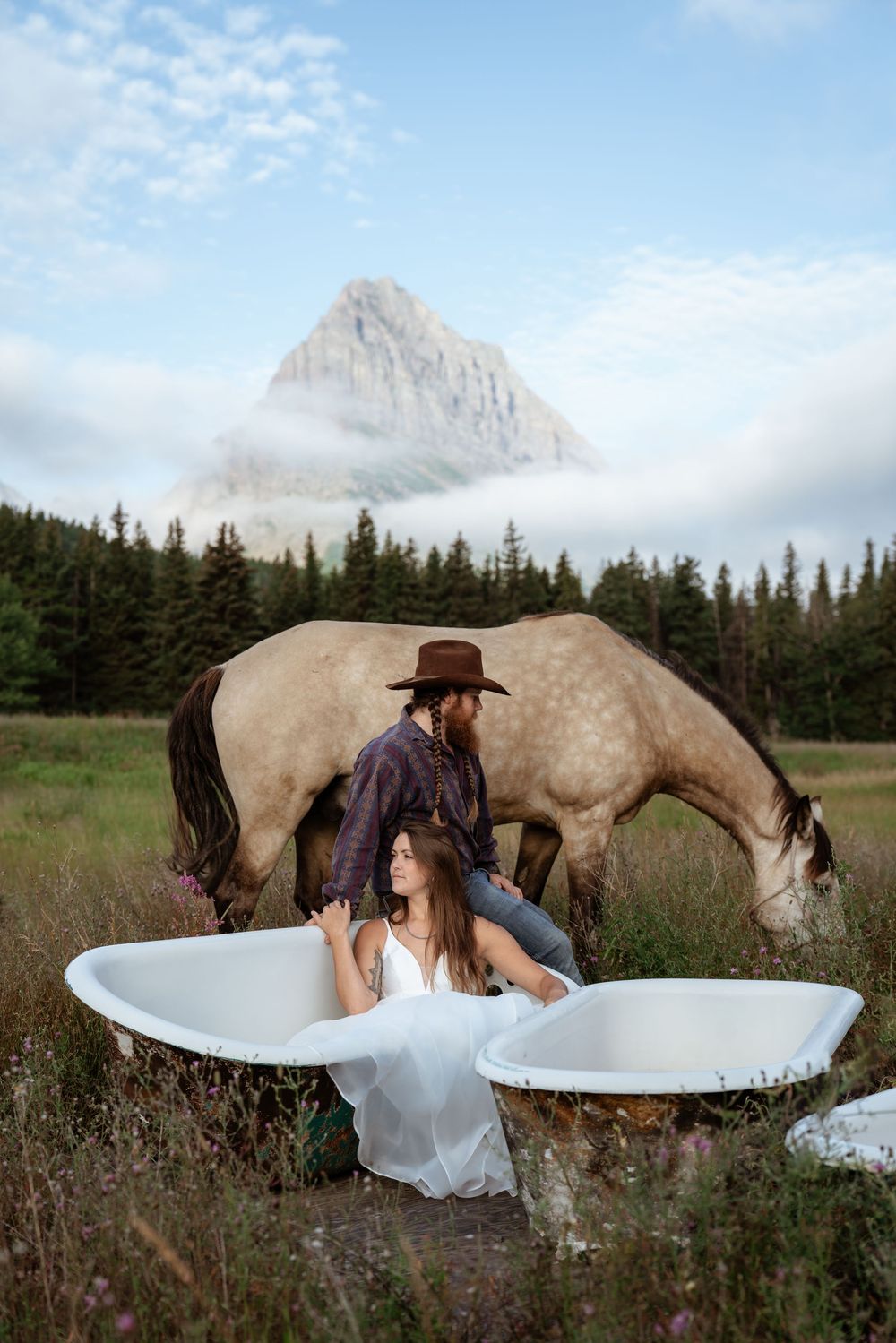 Western bride and groom sit in abandoned bathtubs in large grassy field with buckskin horse and mountain behind them.