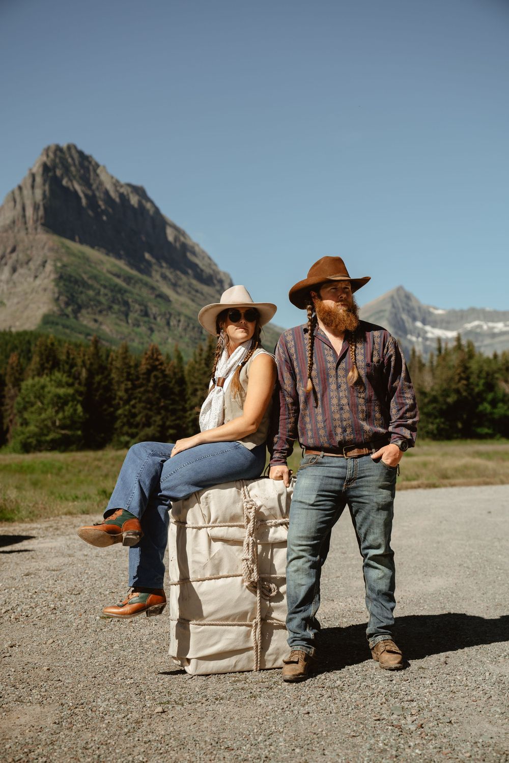 Two people in cowboy hats sit on mantied mule packer load in front of scenic mountain peaks in Glacier National Park.