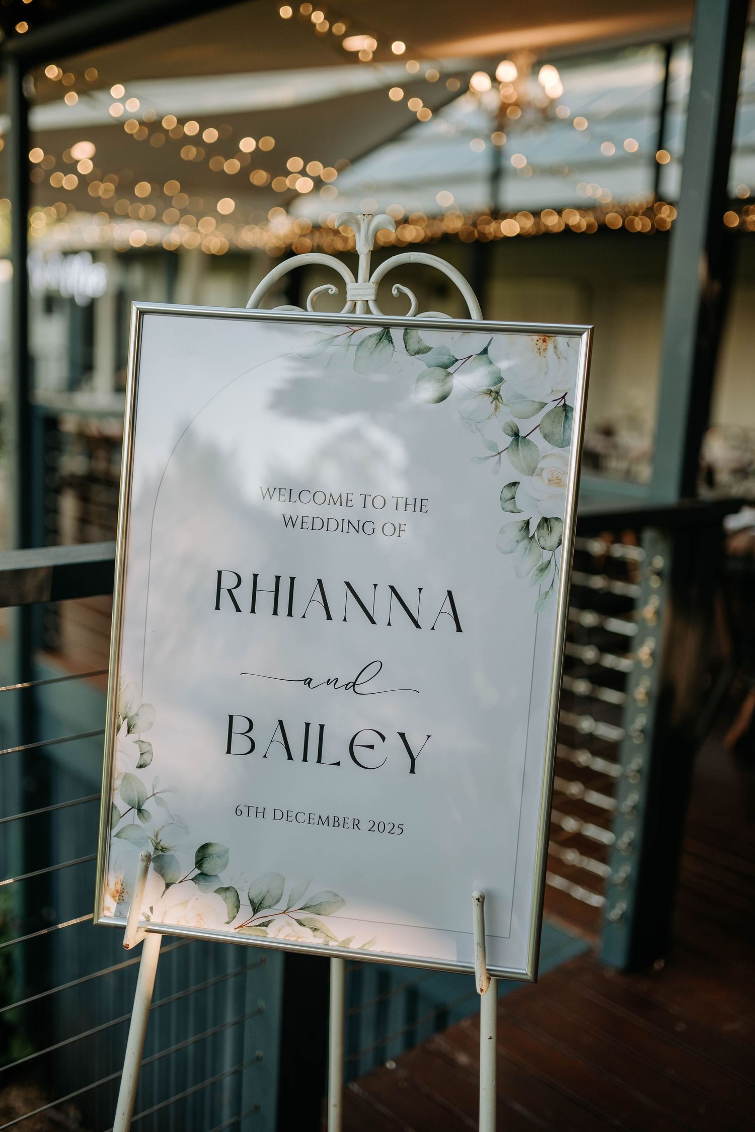 Elegant wedding signage with floral accents and string lights in a romantic venue setting.
