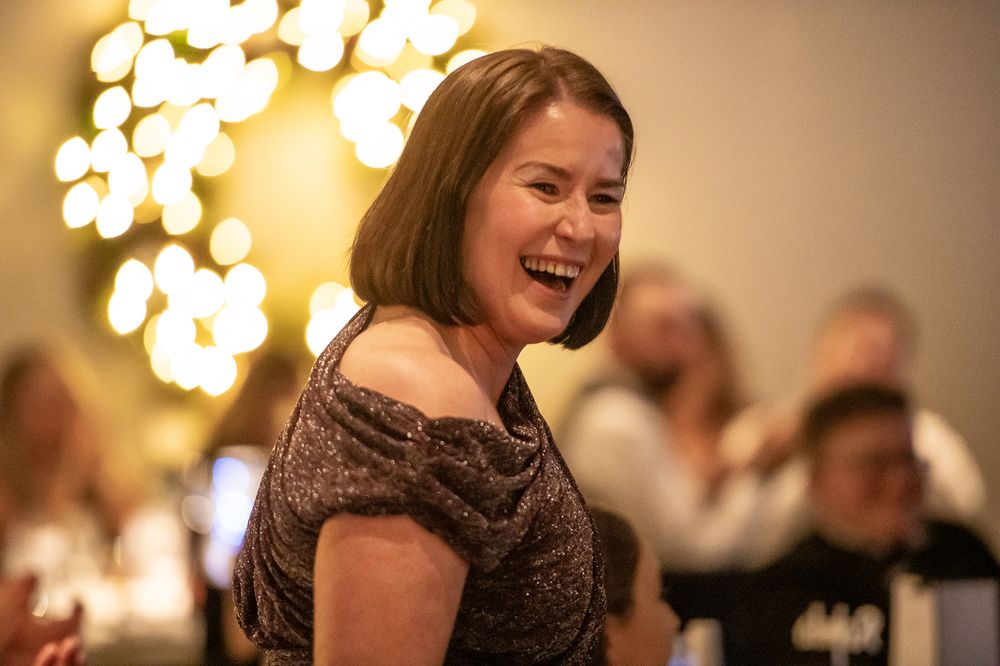 A person in a formal dress smiles and laughs during what appears to be a celebratory event with glowing lights in background.