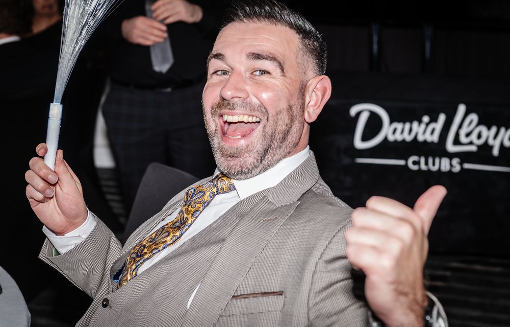 Man in light colored suit smiling and gesturing at a nightclub event.