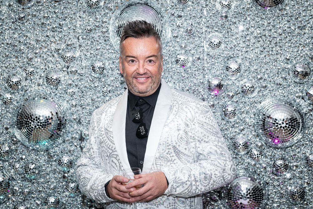 A performer in a sparkly white sequined jacket poses against a glittery silver backdrop with disco ball reflections.