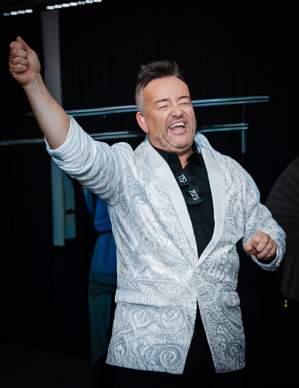 A performer in a white patterned jacket and black shirt gestures enthusiastically during an energetic stage performance.