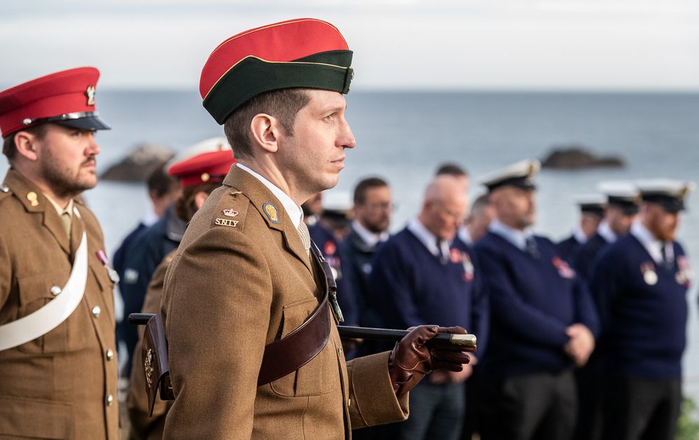 Military personnel in red berets and brown uniforms stand in formation during a ceremony by the ocean.