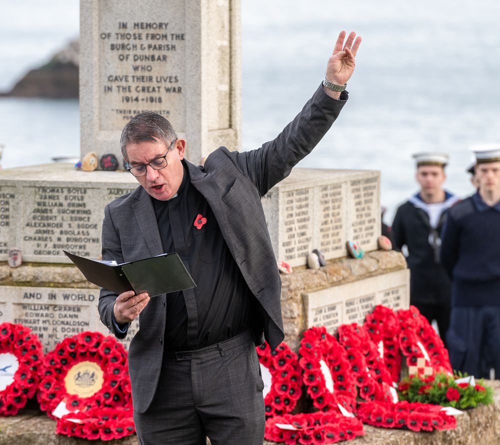 A person stands among bright red poppy wreaths at a memorial service while raising their arm in greeting.