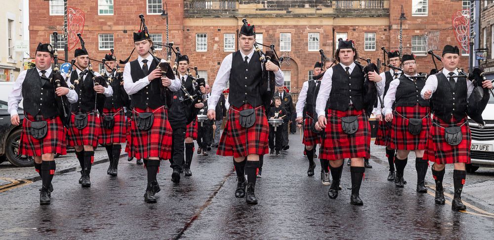 Scottish dancers in red tartan kilts perform a traditional dance routine on a wet cobblestone street.
