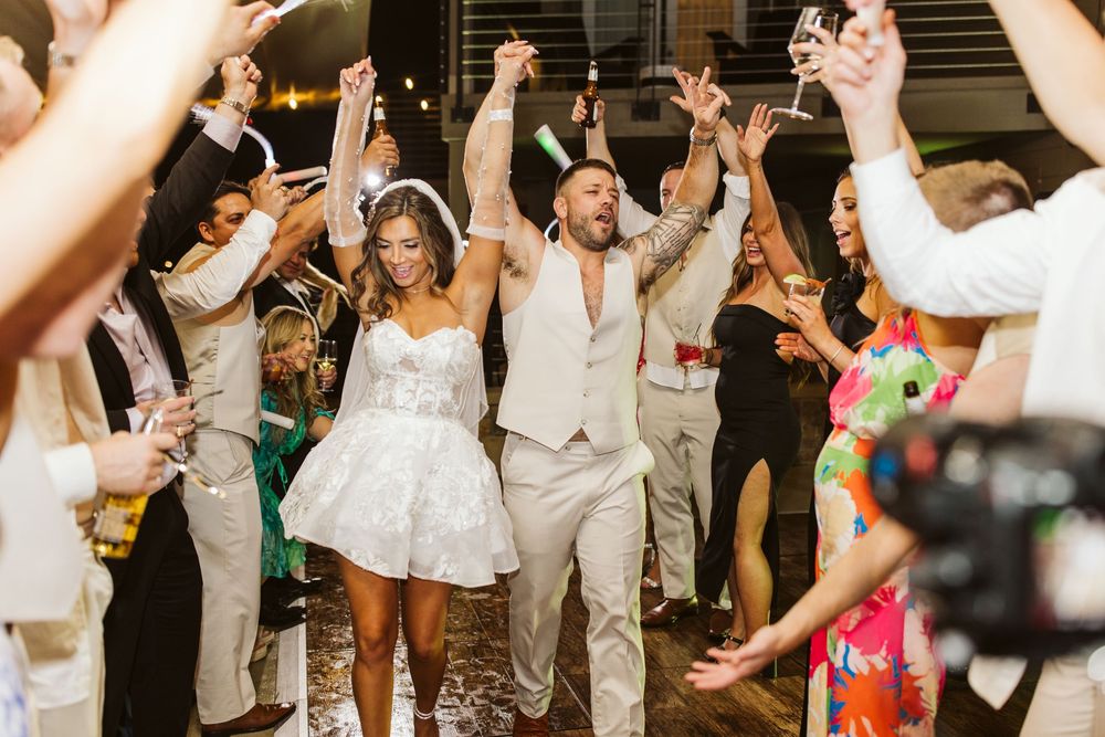 Wedding guests dance and celebrate on a crowded dance floor at a reception party with hands raised in joyful celebration.