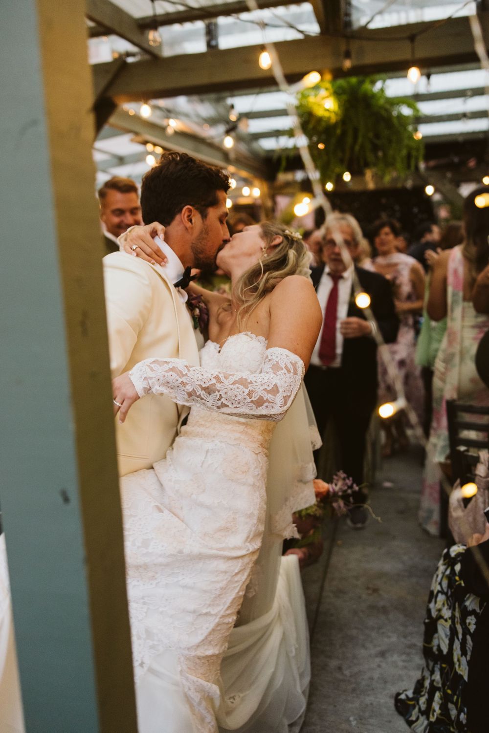 A romantic wedding couple shares their first dance while guests look on at an indoor reception venue with string lights overhead.