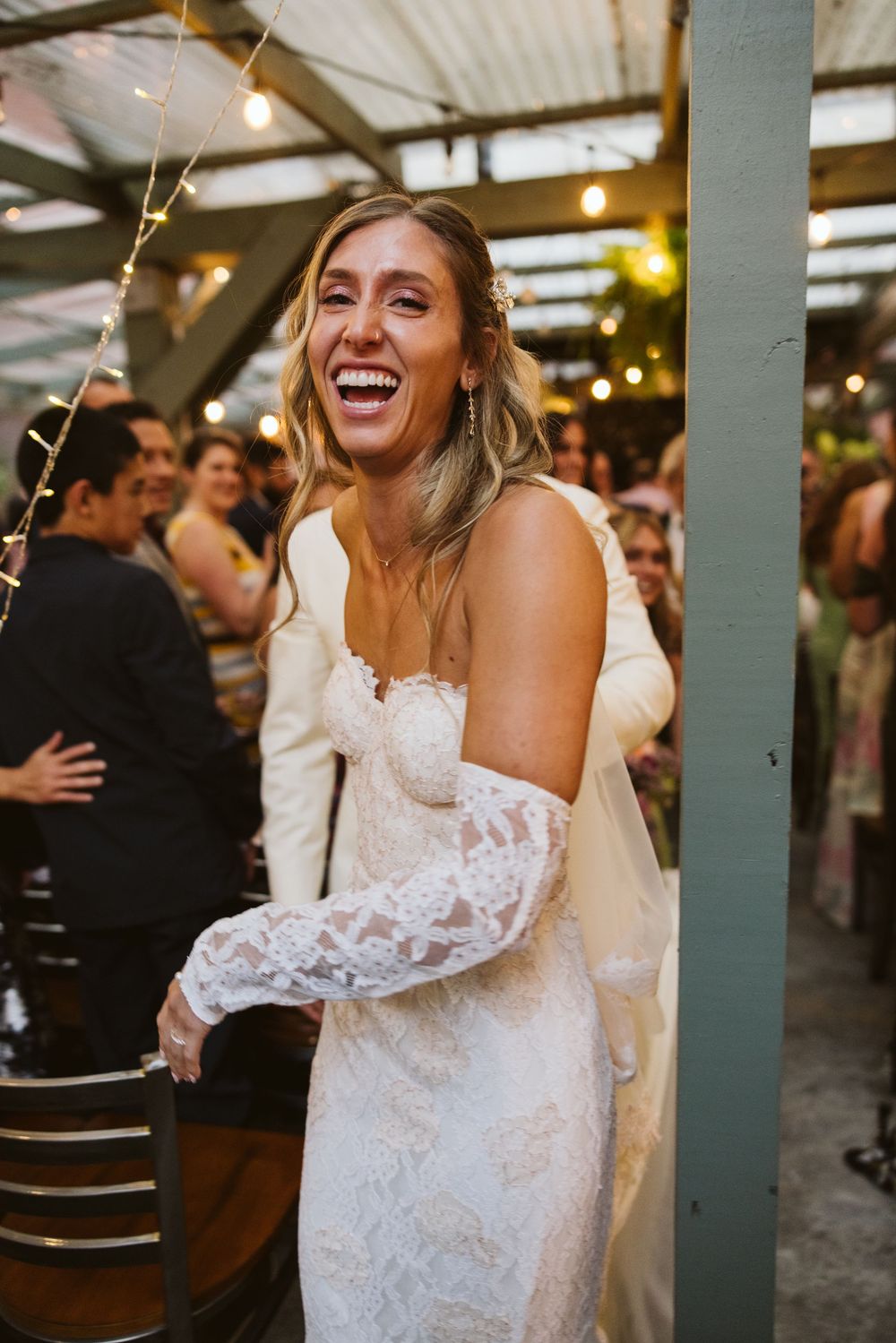 A bride in a white lace wedding dress laughs joyfully during a reception celebration under string lights.