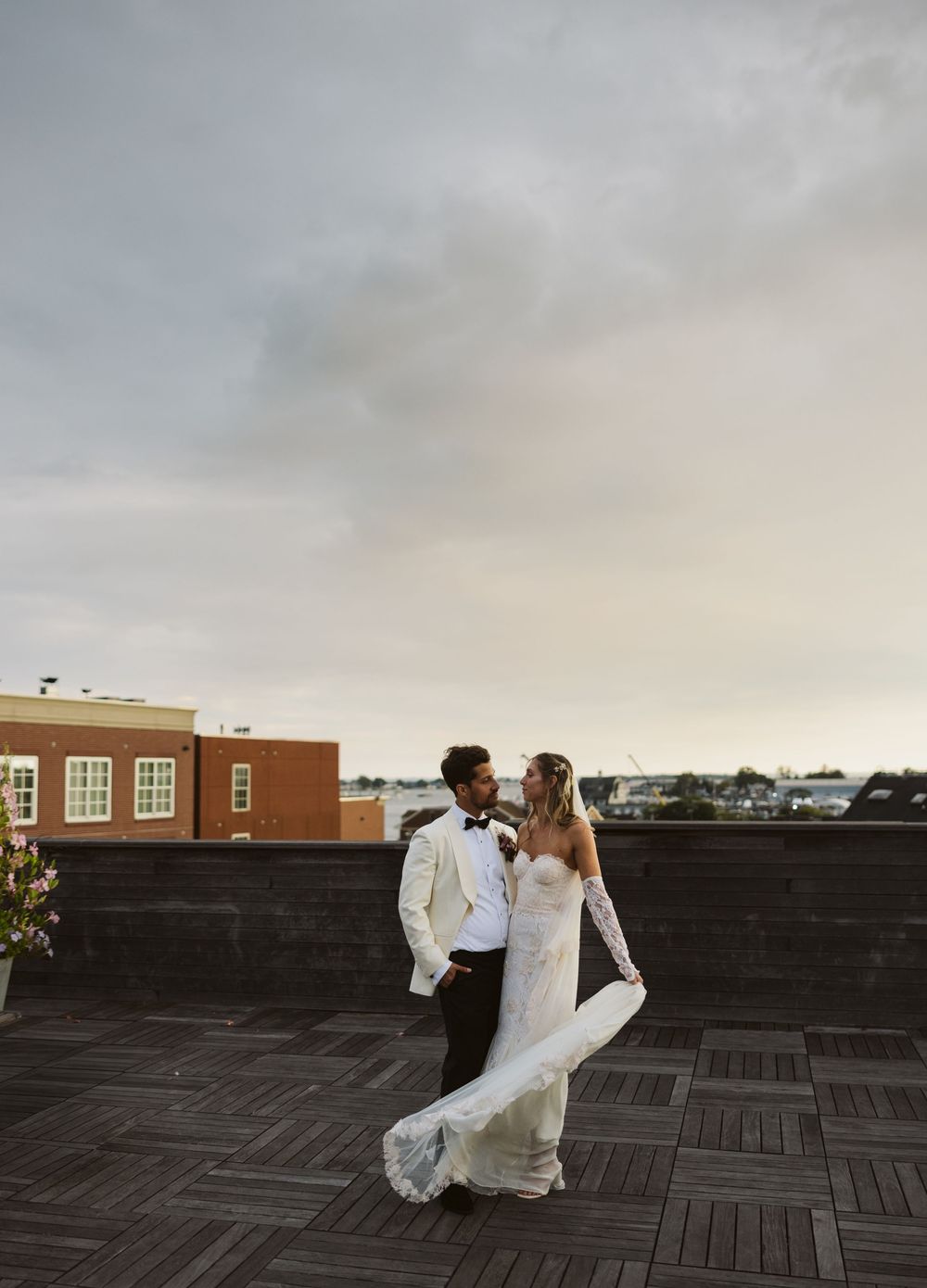 A couple in white wedding attire dances together on a rooftop terrace at sunset.