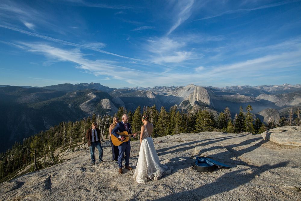 Hikers stand atop Sentinel Dome overlooking Half Dome and Yosemite Valley on a clear sunny day with blue skies.
