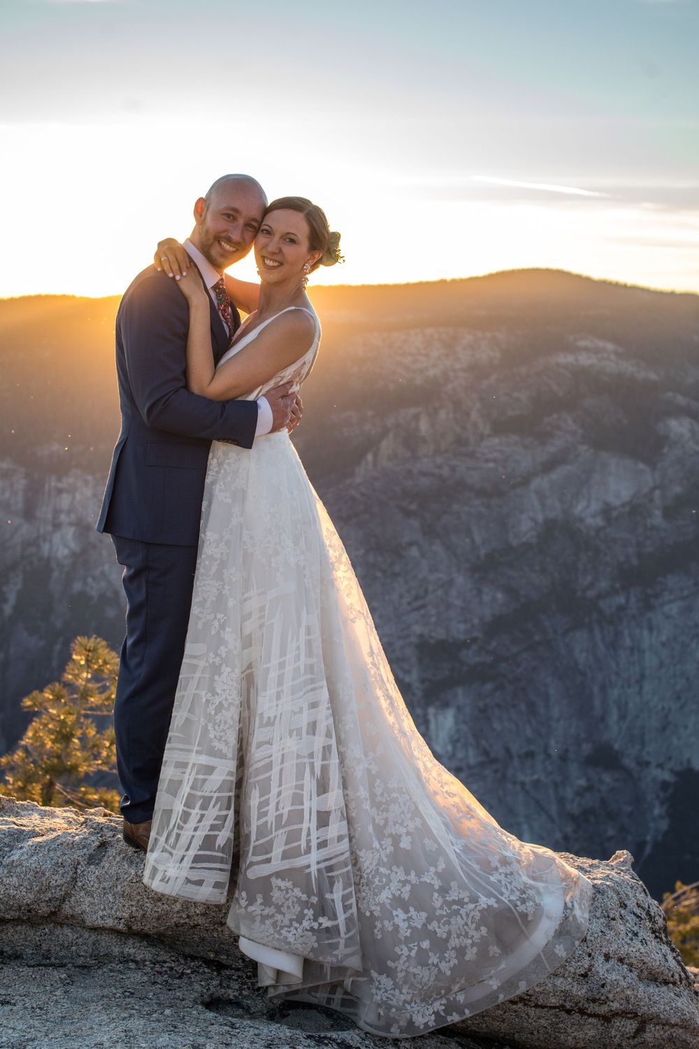 A couple shares a romantic sunset moment on a mountain cliff overlooking dramatic valley views in their wedding attire.