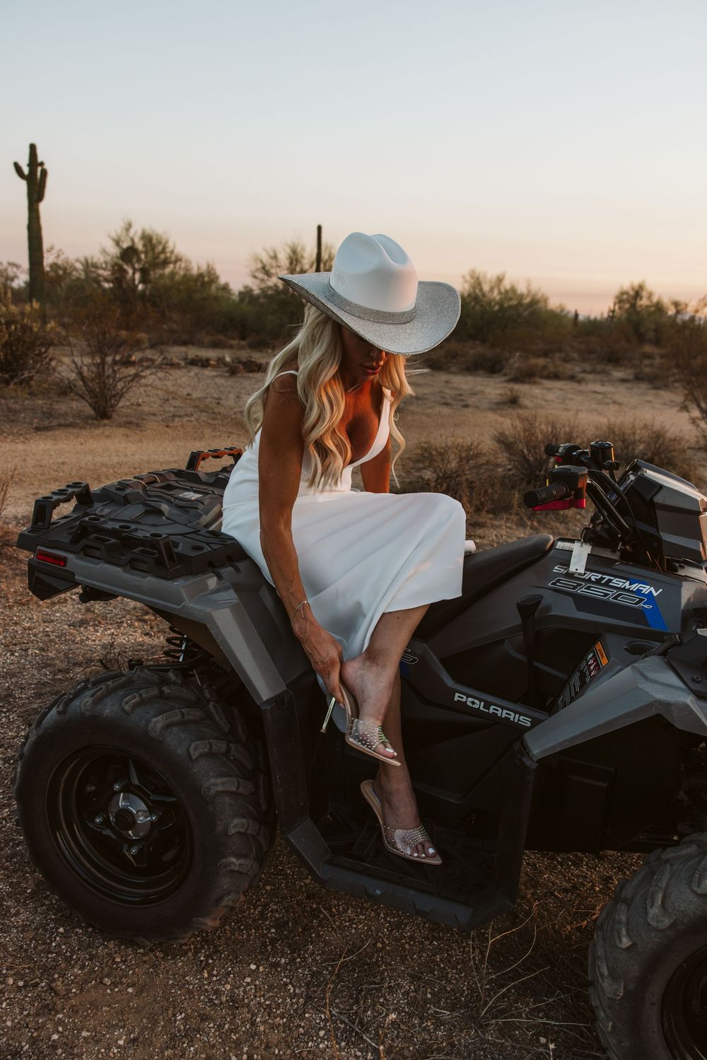 A person in a white dress and cowboy hat sits on a black ATV in the desert.