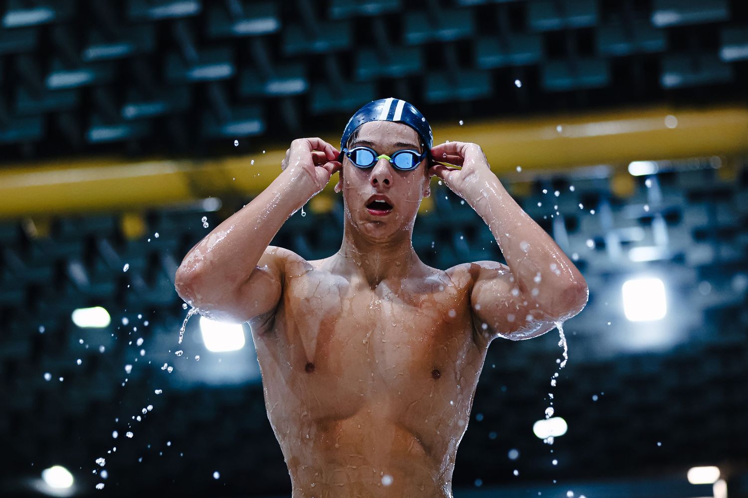 Competitive swimmer adjusts goggles while standing shirtless with water droplets splashing around in dark arena.