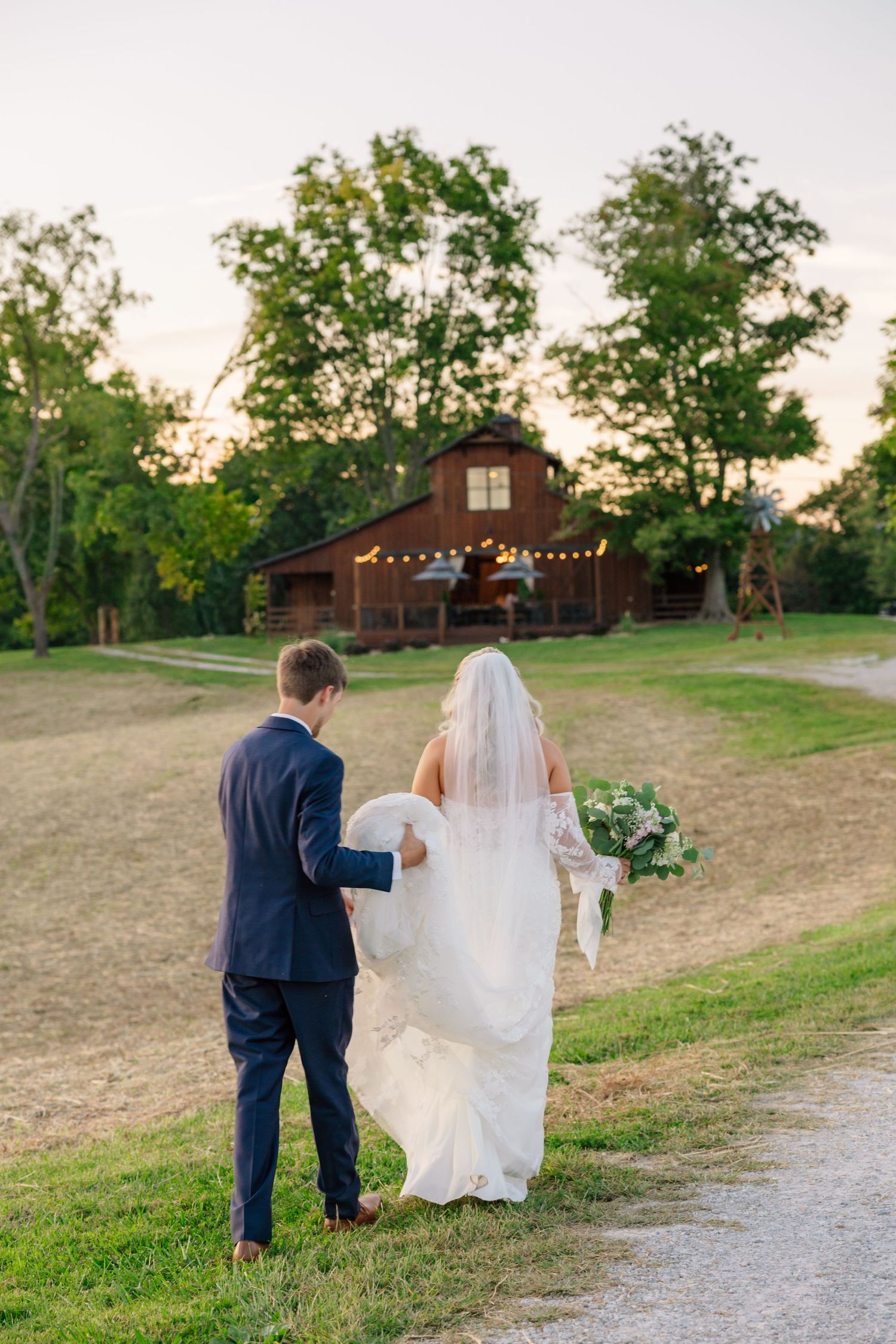 A couple walks down a gravel path towards a rustic barn venue at sunset.