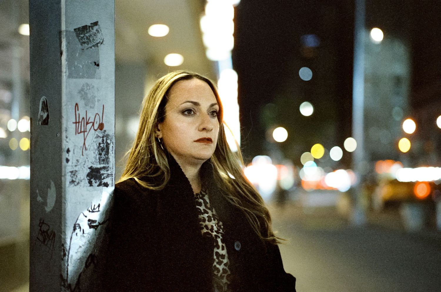 Portrait of someone in a dark coat leaning against a street pole at night with city lights in background.